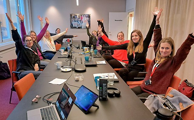 Åtta personer runt ett konferensbord, med armarna uppsträckta åt olika håll.